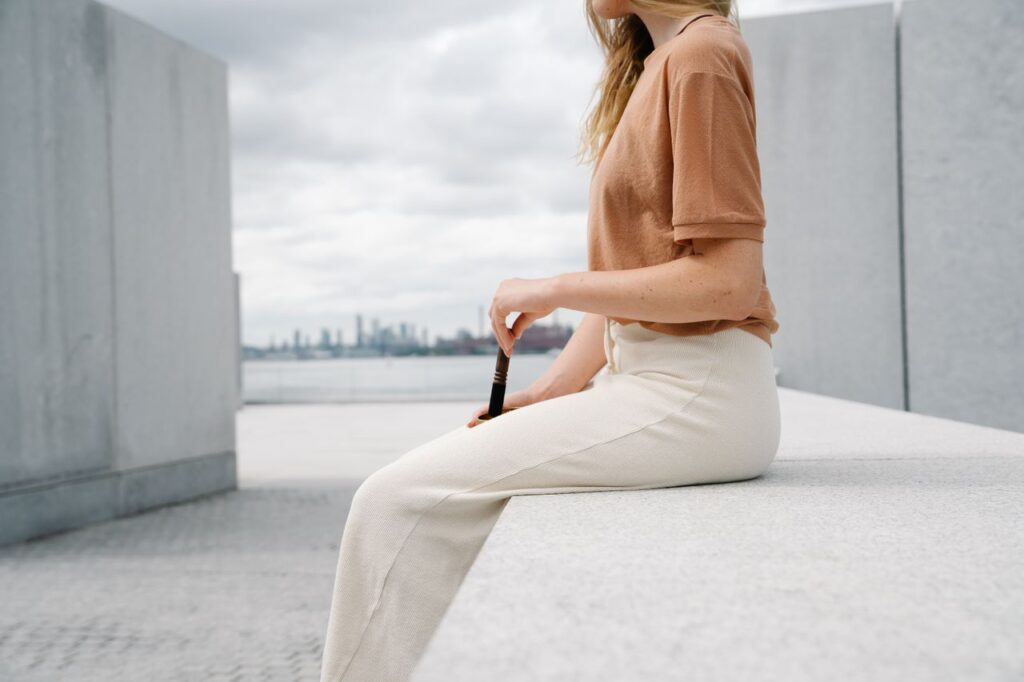 Stylish woman in brown t-shirt seated outdoors on a concrete platform with city skyline view.